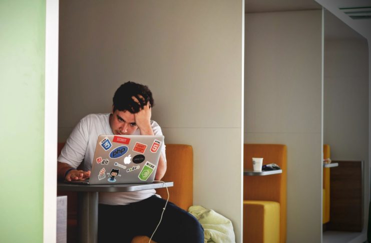 Man sitting in a booth looking at his computer, overwhelmed, with hand on his head. Overcome The Feeling Of Overwhelm